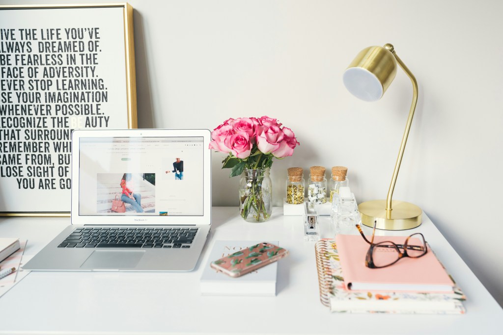 Photo of a silver laptop on a tidy white desk. There is a big motivational sign on the left. To the right is a golden lamp, pair of glasses and some notebooks.