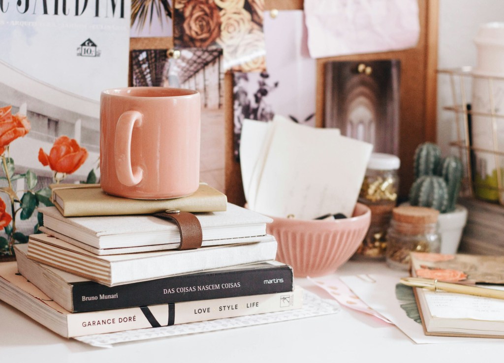 Photo from Unsplash by Ella Jardim Shoes a peach coloured mug sitting on top of a stack of notebooks in a busy, pretty office space.